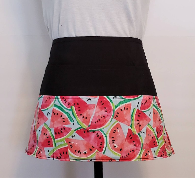 May include: A black apron with a watermelon print. The apron has two pockets on the front and a wide waistband.