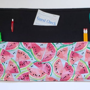 May include: Black fabric organizer with multiple pockets and a watermelon print. The organizer has two long ties on each side. The pockets contain a guest check, scissors, pencils, and markers.