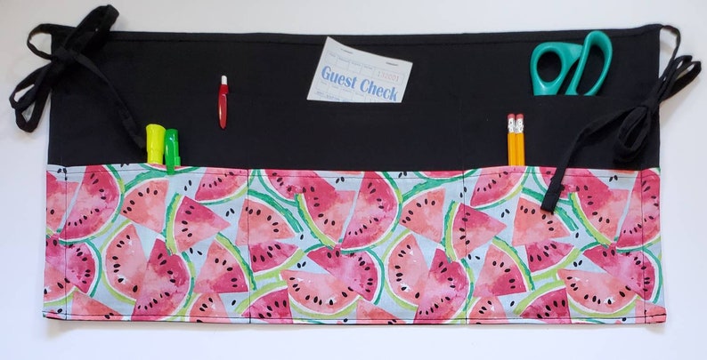 May include: Black fabric organizer with multiple pockets and a watermelon print. The organizer has two long ties on each side. The pockets contain a guest check, scissors, pencils, and markers.