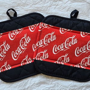 Könnte beinhalten: Zwei schwarze und rote Topflappen mit gesteppter Oberfläche. Die Topflappen haben ein rotes Stoffteil mit dem Coca-Cola-Logo, das in Weiß wiederholt wird.