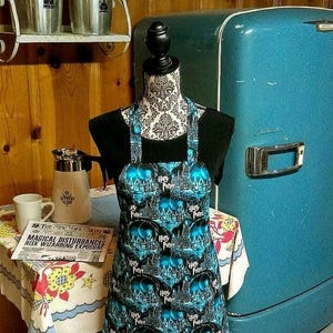 Puede incluir: Un delantal negro con un estampado de Harry Potter azul y blanco. El delantal tiene tirantes ajustables y un bolsillo grande en la parte delantera. El delantal está colgado de un maniquí frente a un refrigerador azul.