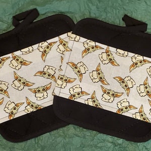 Puede incluir: Dos agarraderas negras con un panel blanco con un patrón repetido de un personaje de dibujos animados con orejas y ojos grandes. Cada agarradera tiene un lazo negro para colgar.