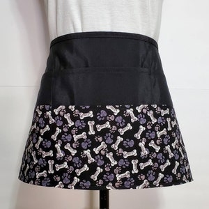 K&ouml;nnte beinhalten: Schwarze Sch&uuml;rze mit einer gro&szlig;en Tasche und einem unteren Rand mit einem wei&szlig;en und violetten Pfoten- und Knochenmuster. Das Muster wiederholt das Wort "Knochen" durchgehend.