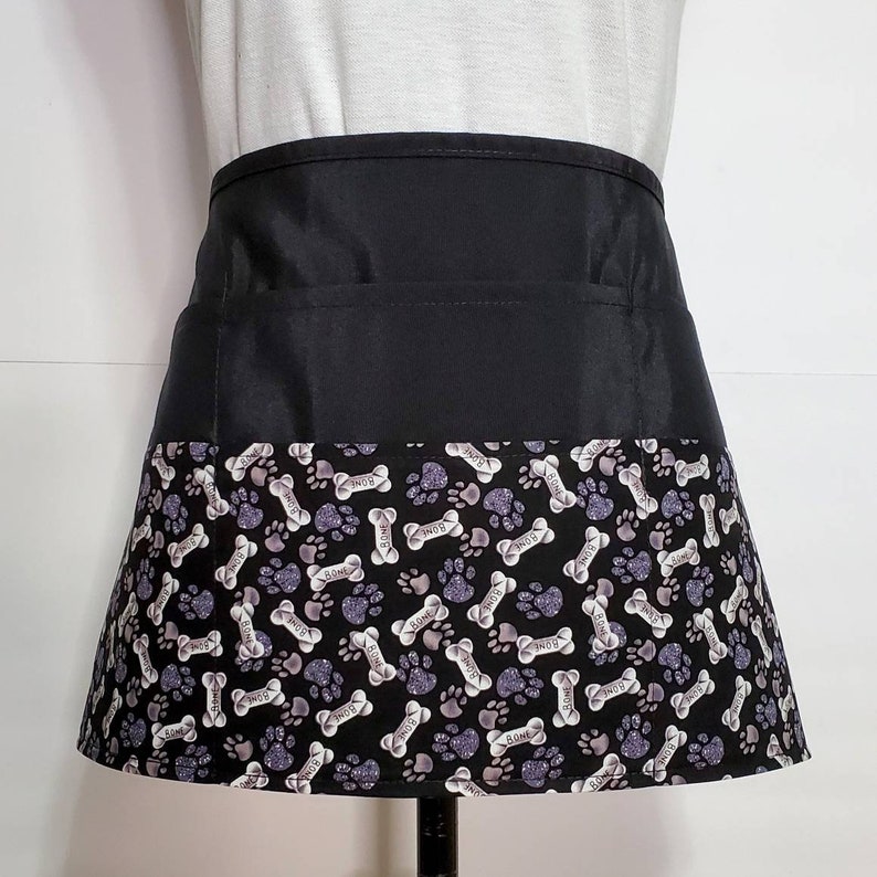 K&ouml;nnte beinhalten: Schwarze Sch&uuml;rze mit einer gro&szlig;en Tasche und einem unteren Rand mit einem wei&szlig;en und violetten Pfoten- und Knochenmuster. Das Muster wiederholt das Wort "Knochen" durchgehend.