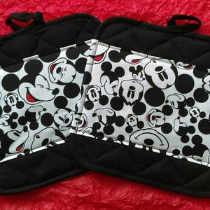 Puede incluir: Dos agarraderas de olla negras y blancas con un patrón de Mickey Mouse. Las agarraderas tienen un respaldo acolchado negro y un frente de tela blanca con lunares negros y caras de Mickey Mouse.