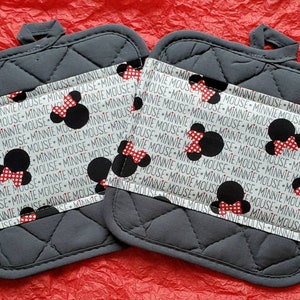 May include: Two gray quilted pot holders with a white fabric band featuring a repeating pattern of Minnie Mouse heads and the text "Minnie Mouse".