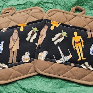 May include: Two brown quilted pot holders with a black fabric panel featuring a repeating pattern of Star Wars characters including Chewbacca, Yoda, R2-D2, C-3PO, and BB-8.
