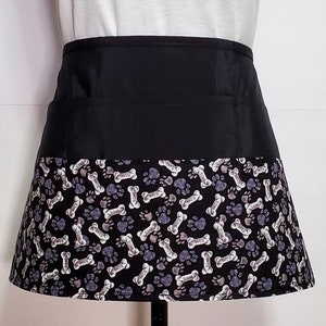 K&ouml;nnte beinhalten: Schwarze Sch&uuml;rze mit schwarzem Bund und einem unteren Teil mit einem wei&szlig;en und grauen Pfoten- und Knochenmuster. Das Muster wiederholt das Wort "Bone" durchgehend.