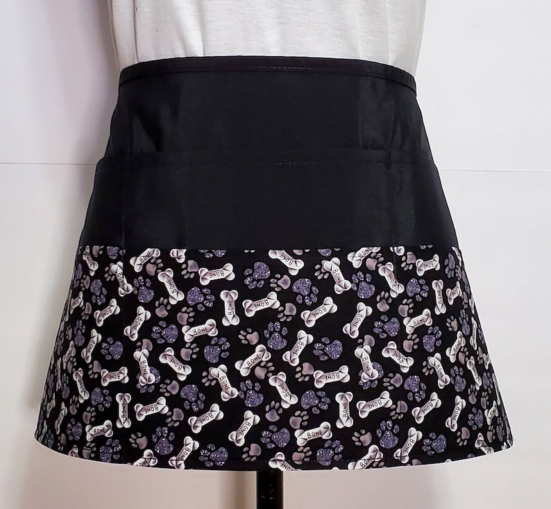 K&ouml;nnte beinhalten: Schwarze Sch&uuml;rze mit schwarzem Bund und einem unteren Teil mit einem wei&szlig;en und grauen Pfoten- und Knochenmuster. Das Muster wiederholt das Wort "Bone" durchgehend.