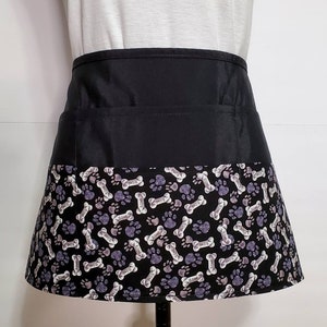 K&ouml;nnte beinhalten: Schwarze Sch&uuml;rze mit einer gro&szlig;en Tasche und einem unteren Rand mit einem wei&szlig;en und grauen Pfoten- und Knochenmuster, auf dem das Wort "Bone" wiederholt vorkommt.