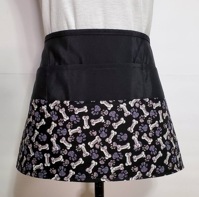 K&ouml;nnte beinhalten: Schwarze Sch&uuml;rze mit einer gro&szlig;en Tasche und einem unteren Rand mit einem wei&szlig;en und grauen Pfoten- und Knochenmuster, auf dem das Wort "Bone" wiederholt vorkommt.