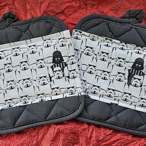 Può includere: Un paio di guanti da forno imbottiti neri con un motivo bianco e nero di stormtrooper e Darth Vader. I guanti sono realizzati in tessuto imbottito e hanno un anello per appenderli.