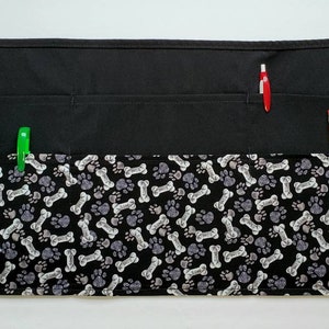 K&ouml;nnte beinhalten: Schwarze Sch&uuml;rze mit einem Pfoten- und Knochenmuster. Die Sch&uuml;rze hat zwei gro&szlig;e Taschen und zwei kleinere Taschen. In den Taschen befinden sich zwei Bleistifte und ein Stift.