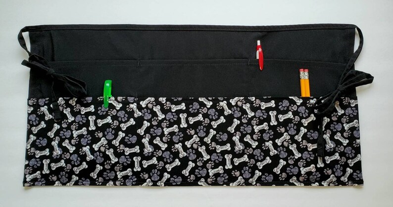 K&ouml;nnte beinhalten: Schwarze Sch&uuml;rze mit einem Pfoten- und Knochenmuster. Die Sch&uuml;rze hat zwei gro&szlig;e Taschen und zwei kleinere Taschen. In den Taschen befinden sich zwei Bleistifte und ein Stift.