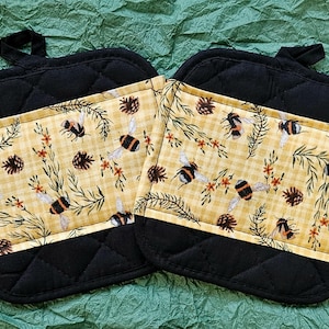 May include: Two black quilted oven mitts with a yellow and white patterned pocket featuring bees, pinecones, and sprigs. The mitts have a loop for hanging. The background is a textured green surface.