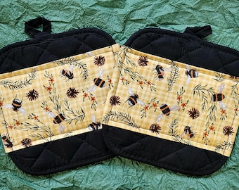 May include: Two black quilted oven mitts with a yellow and white patterned pocket featuring bees, pinecones, and sprigs. The mitts have a loop for hanging. The background is a textured green surface.