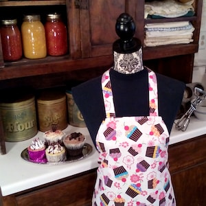 May include: A white apron with a colorful cupcake pattern. The apron has adjustable straps and a large pocket in the front. The apron is hanging on a black mannequin.