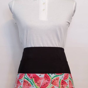 May include: Black apron with a watermelon print. The apron has a black waistband and a pocket on the front.