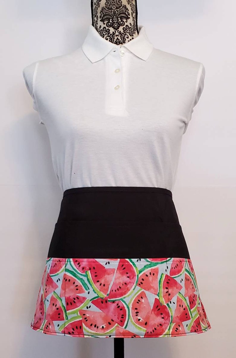 May include: Black apron with a watermelon print. The apron has a black waistband and a pocket on the front.