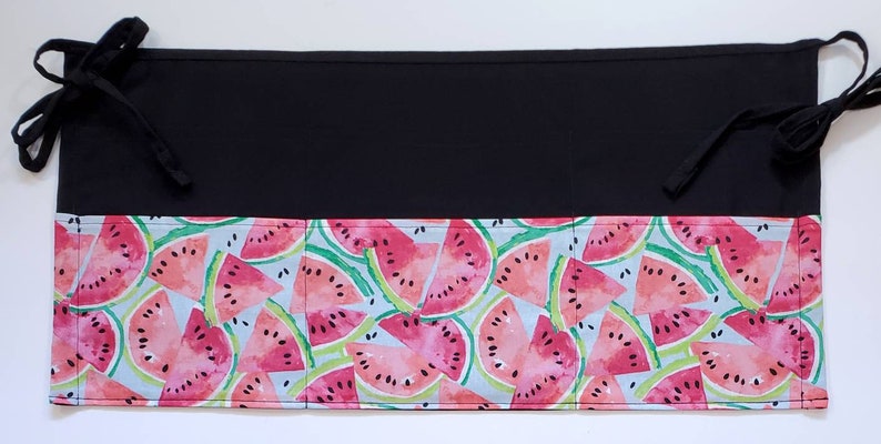May include: Black fabric apron with a watermelon print. The apron has three pockets and ties at the waist.