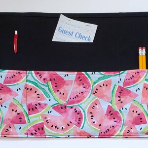 May include: Black fabric apron with multiple pockets, featuring a watermelon print. The apron has two black ties on each side. The top pocket has a guest check and a red pen. The other pocket has two yellow pencils and a pair of green scissors.