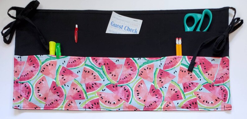 May include: Black fabric apron with multiple pockets, featuring a watermelon print. The apron has two black ties on each side. The top pocket has a guest check and a red pen. The other pocket has two yellow pencils and a pair of green scissors.
