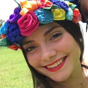May include: A woman wearing a colorful felt flower crown. The crown is made of felt flowers in shades of pink, blue, yellow, orange, and green. The woman is smiling and has long dark hair.