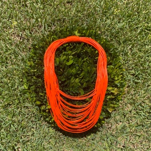 May include: A bright orange layered necklace made of thin, flexible cord. The necklace is laid out on a bed of green grass.