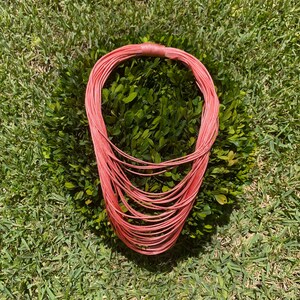 May include: A coral-colored multi-strand necklace made of thin cords. The necklace is laid out in a circle on a bed of green grass.