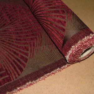May include: A roll of burgundy fabric with a brown geometric pattern. The fabric has a textured surface and a fringed edge.