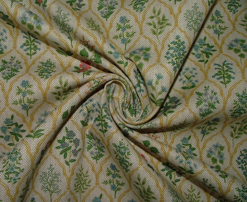 May include: Close-up of a beige fabric with a repeating floral pattern. The design features green, blue, and pink floral elements within a gold trellis-like framework. The fabric is gathered, showing its texture and folds.