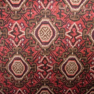 May include: Close-up of a patterned fabric with a repeating geometric design. The pattern features a mix of red, brown, and cream colors. The design includes diamond and oval shapes, creating a textured appearance.