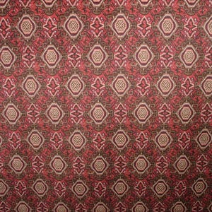 May include: Close-up of a patterned fabric with a repeating geometric design. The pattern features diamond and circular shapes in shades of red, brown, and beige. The fabric has a textured appearance, suggesting a woven material.