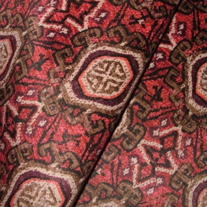 May include: Close-up of a folded fabric with a detailed, repeating pattern. The design features geometric shapes and stylized floral elements in shades of red, brown, and cream. The fabric has a textured appearance.