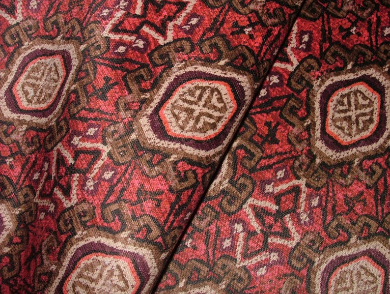 May include: Close-up of a folded fabric with a detailed, repeating pattern. The design features geometric shapes and stylized floral elements in shades of red, brown, and cream. The fabric has a textured appearance.