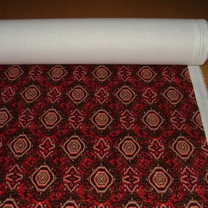 May include: A roll of fabric with a repeating geometric pattern in red, brown, and tan. The pattern features octagonal shapes with intricate designs. The fabric is partially unrolled, revealing a white backing.