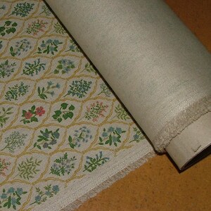 May include: A roll of beige fabric with a floral pattern. The pattern features small flowers in pink, blue, and green, set within a yellow honeycomb design. The fabric is partially unrolled, revealing the plain beige backing.