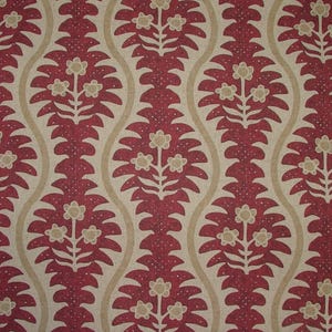 May include: A close-up of a fabric with a repeating pattern of stylized floral motifs. The design features a deep red color with cream-coloured accents and a beige background. The floral elements are symmetrical and detailed.