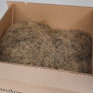May include: A cardboard box filled with brown packing material. The box is open, revealing a large quantity of dried, fibrous material, likely used for cushioning or decorative purposes. The box is positioned at an angle, with the contents visible.