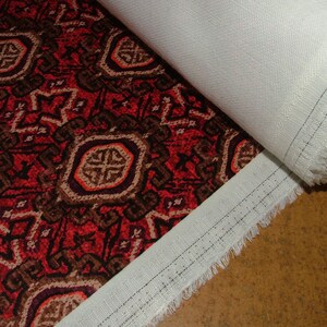 May include: A roll of fabric featuring a detailed, repeating pattern in shades of red, brown, and beige. The design includes geometric shapes and floral elements. The fabric is partially rolled, revealing a white backing.