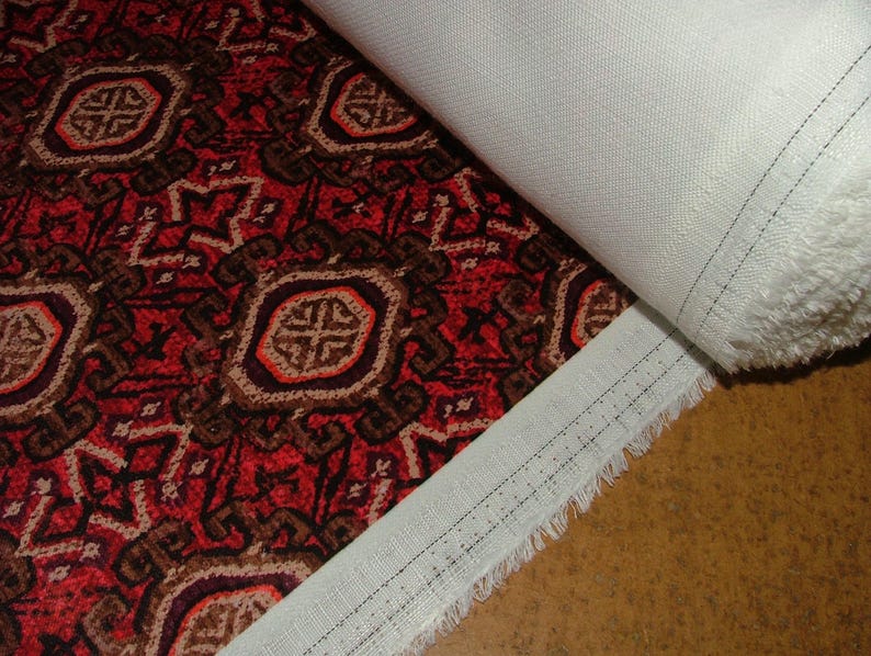 May include: A roll of fabric featuring a detailed, repeating pattern in shades of red, brown, and beige. The design includes geometric shapes and floral elements. The fabric is partially rolled, revealing a white backing.