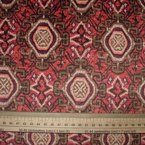 May include: Close-up of a patterned fabric with a ruler. The fabric features a repeating design of geometric shapes in shades of red, brown, and cream. The ruler shows measurements in inches and centimeters.