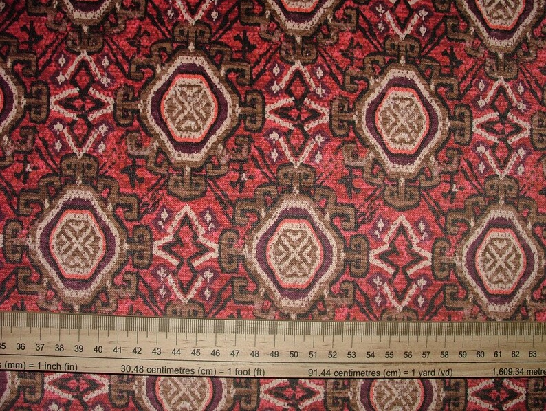 May include: Close-up of a patterned fabric with a ruler. The fabric features a repeating design of geometric shapes in shades of red, brown, and cream. The ruler shows measurements in inches and centimeters.