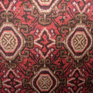 May include: Close-up of a patterned textile with a repeating geometric design. The pattern features a mix of red, brown, and cream colors. The design includes stylized shapes and a central medallion-like element, creating a textured, ornate appearance.