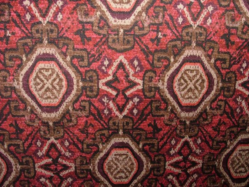 May include: Close-up of a patterned textile with a repeating geometric design. The pattern features a mix of red, brown, and cream colors. The design includes stylized shapes and a central medallion-like element, creating a textured, ornate appearance.