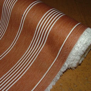 May include: A rolled fabric with a brown base and white vertical stripes. The material has a textured weave and a fringed edge. The fabric is suitable for upholstery or home decor, with a visible roll of the material.