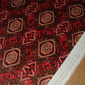 May include: Close-up of a patterned fabric with a red background and repeating geometric designs in brown, beige, and orange. A white fabric edge with a stitched seam is visible on the right side, suggesting a textile or upholstery material.