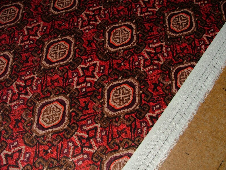 May include: Close-up of a patterned fabric with a red background and repeating geometric designs in brown, beige, and orange. A white fabric edge with a stitched seam is visible on the right side, suggesting a textile or upholstery material.