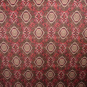 May include: Close-up of a patterned fabric with a repeating geometric design. The pattern features diamond and oval shapes in shades of red, brown, and beige. The fabric has a textured appearance, suggesting a woven or embroidered construction.