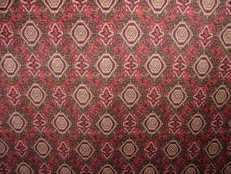 May include: Close-up of a patterned fabric with a repeating geometric design. The pattern features diamond and oval shapes in shades of red, brown, and beige. The fabric has a textured appearance, suggesting a woven or embroidered construction.
